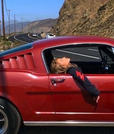 a woman sticking her head out the window of a red car with mountains in the background