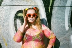 a woman wearing sunglasses standing in front of a graffiti wall with her hand on her head