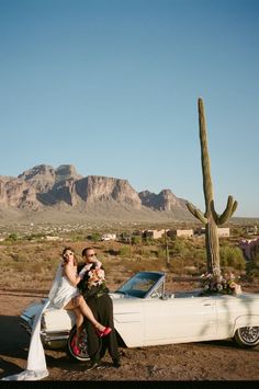 a bride and groom sitting on the back of a white car in front of a cactus