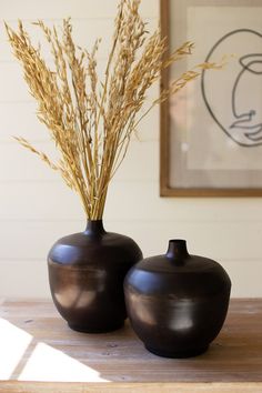 two black vases sitting on top of a wooden table next to each other with dry grass in them