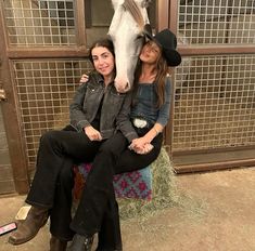 two women sitting on a stool with a horse in the background and hay behind them