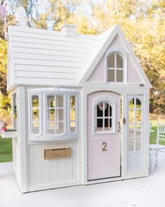 a white doll house with pink doors and windows on the front porch, next to a bench