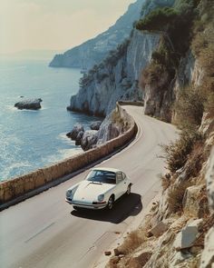 a white car is driving down the road by the water's edge, with cliffs in the background