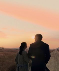 a man and woman walking down a dirt road at sunset with the sun setting behind them