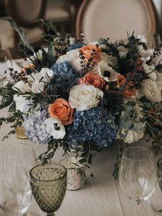 an arrangement of flowers and greenery is sitting on a table with wineglasses