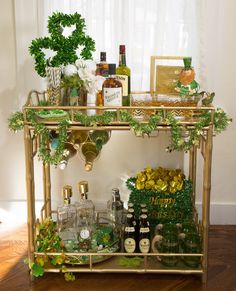 a gold bar cart with bottles and glasses on it, surrounded by greenery in front of a window