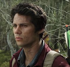 a close up of a person in a forest with trees and bushes behind him, looking at the camera