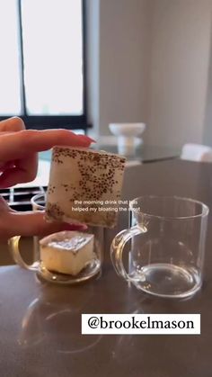 someone holding a piece of cake next to a glass cup on a table with other items