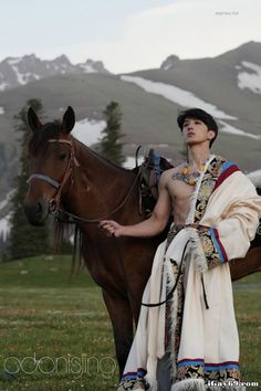 a man in native garb standing next to a horse with mountains in the background