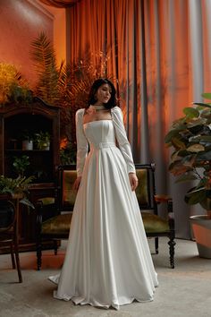 a woman in a white wedding dress standing next to a chair and potted plant