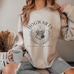 a woman wearing a harry potter sweatshirt sitting on a chair holding a coffee mug in her hand