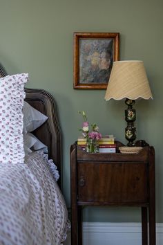 a nightstand with flowers and books on it next to a bed in a room that has green walls