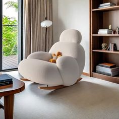 a white chair sitting in front of a window next to a book shelf filled with books