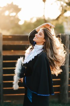 a woman with long hair wearing a black outfit and white fur stoler, standing in front of a fence