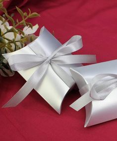 two white gift boxes with ribbons tied around them on a red tablecloth next to flowers