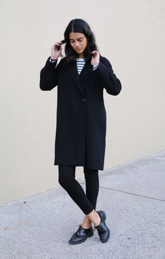 a woman in black coat and striped shirt standing on the sidewalk with her hands behind her head