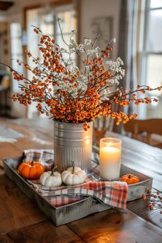 a table topped with a metal container filled with oranges and white pumpkins next to a candle