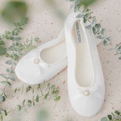 two white shoes with bows and pearls on them sitting next to some green leaves in the sand