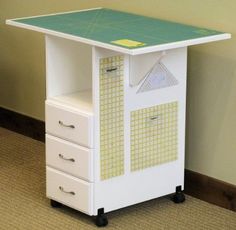 a small white cart with three drawers and a green top on wheels in front of a wall