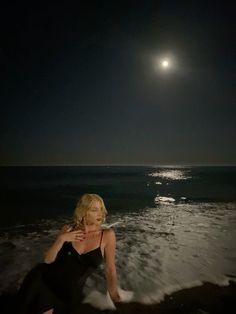 a woman sitting on the beach in front of the ocean at night with full moon