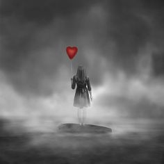 a woman holding a red heart balloon standing on a rock in the middle of water