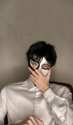 a man wearing a white shirt and black mask covering his face with one hand while sitting in a chair