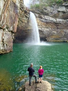 two people standing on rocks in front of a waterfall with green water and cliffs behind them