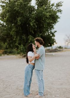 a man and woman standing next to each other in the sand with trees behind them