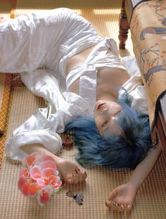 a woman with blue hair laying on the floor next to a bed and flower in her hand