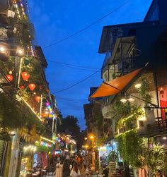 people are walking down an alley way at night with lights on the buildings and plants