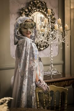 a woman wearing a mask standing in front of a chandelier with candles on it