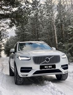 the front end of a white volvo suv driving on snow - covered road with trees in background