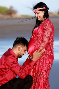 a man kneeling down next to a pregnant woman in a red dress on the beach