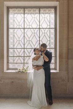 a bride and groom standing in front of a window with their arms around each other