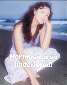 a woman sitting on top of a sandy beach next to the ocean with her hand under her chin