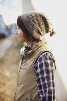 a woman wearing a hat and vest standing next to a white wall on the street