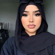 a woman wearing a black hijab and dark makeup looks at the camera with her eyes closed