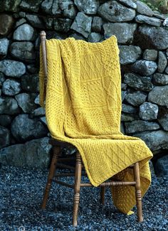 a yellow blanket sitting on top of a wooden chair next to a stone wall with rocks in the background