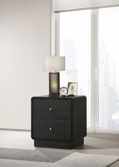 a black night stand with two drawers and a lamp on it in front of a window