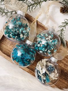 three glass ornaments with blue and white decorations on a wooden tray next to pine branches