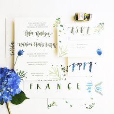 blue flowers and greenery are on top of the wedding stationery, along with some calligraphy