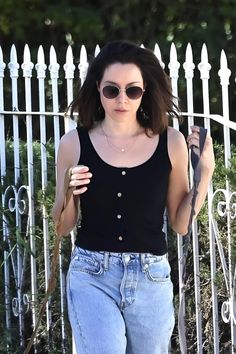 a woman in black shirt and jeans holding an umbrella next to a white picket fence