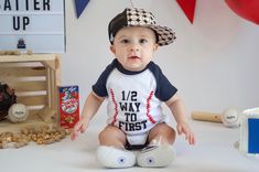 a baby sitting on the ground wearing a baseball shirt and hat with his feet crossed