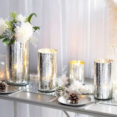 three silver vases filled with white flowers on top of a metal table next to candles