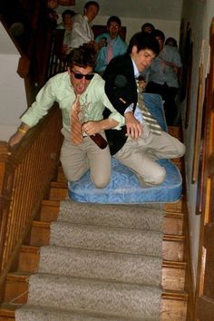 two men sitting on top of a blue cushion in front of stairs with people standing behind them