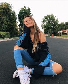 a young woman sitting on the ground with her legs crossed and wearing ripped denims