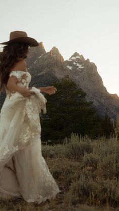 a woman in a white dress and cowboy hat is walking through the grass with mountains in the background