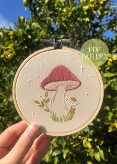 a hand holding up a small embroidery hoop with a mushroom and daisies on it