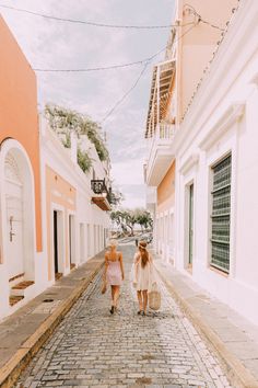 two women walking down the street in front of pink and white buildings, one carrying a bag