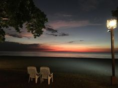 two lawn chairs sitting on top of a grass covered field next to the ocean at night